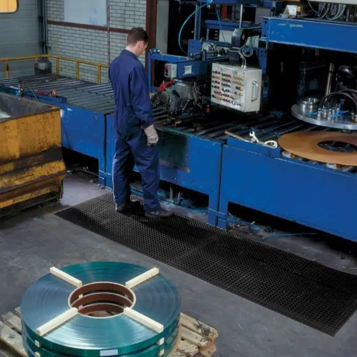Operador em linha de produção metalúrgica utilizando tapete industrial ergonomico antifadiga nr17 para proteção ergonômica em frente a maquinário pesado.